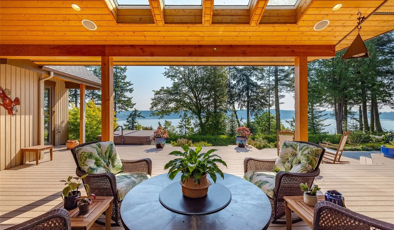 A covered deck with a warm timber ceiling, skylights and exposed beams, looking out past wicker armchairs to a lawn, tall evergreens and open water beyond